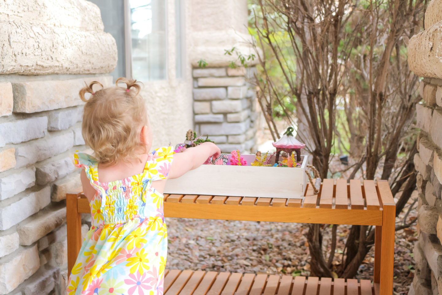 Toddler girl in yellow floral dress standing by her fairy garden