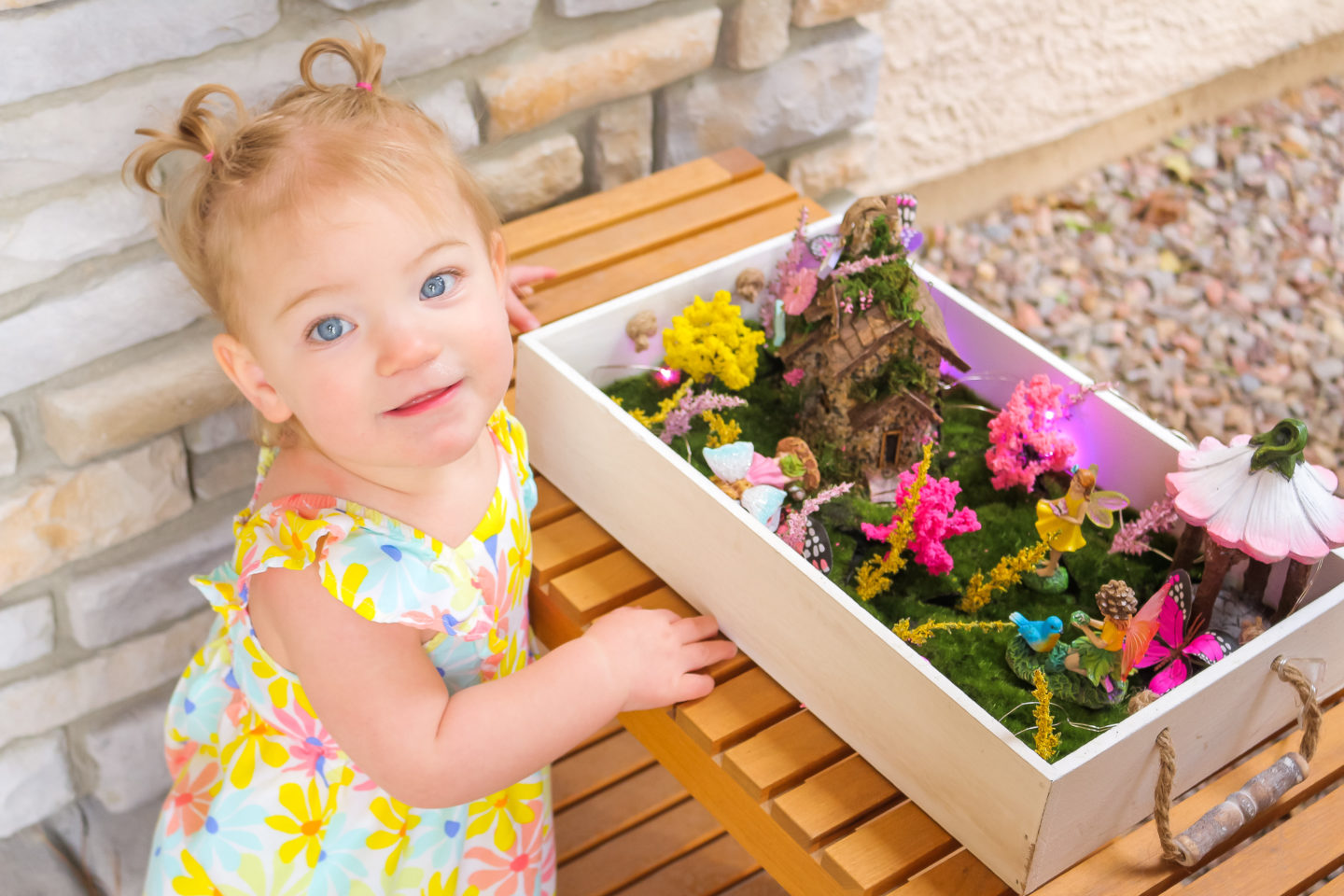 Toddler girl in yellow floral dress standing by her fairy garden